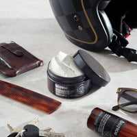 Bullfrog Strong Adhesion Matte Wax, geöffnete Dose mit weißem Wax im Flatlay mit Helm, Kamm und Sonnenbrille