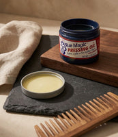 Blue Magic Pressing Oil - offener Tiegel auf Schiefer mit Holzkamm, das gelbe Öl sichtbar